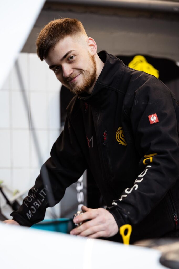Mann mit ernstem Blick in schwarzer Jacke mit gelben Logos am Auto